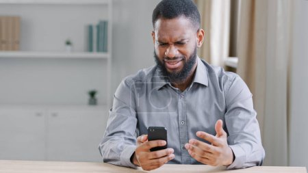 Foto de Angry African American ethnic bearded man boss businessman with mobile phone smartphone mistake failure reading bad news refuse notice mad with business trouble problem sitting at table in office - Imagen libre de derechos