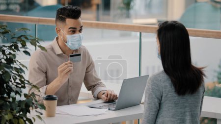 Foto de Man Hispanic bank worker in medical face mask consulting unrecognizable client woman about financial service debit easy payment banking account consumer loan finance saving giving credit card to girl - Imagen libre de derechos