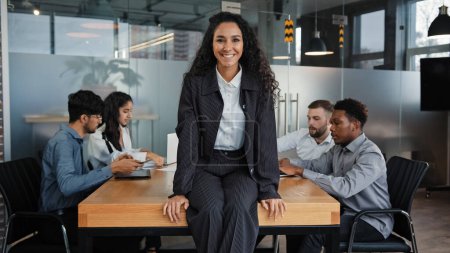 Foto de Satisfied smiling female leader boss woman company CEO businesswoman sitting on table in office smile to camera background of multiracial coworkers colleagues teamwork project startup analyze work - Imagen libre de derechos