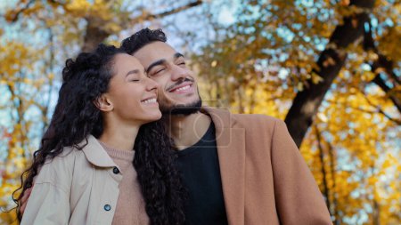 Foto de Ethnic multiracial married couple in autumn park happy smiling Caucasian woman with Hispanic man outdoors guy and girl boyfriend and girlfriend look into distance dreaming together enjoy date outdoors - Imagen libre de derechos