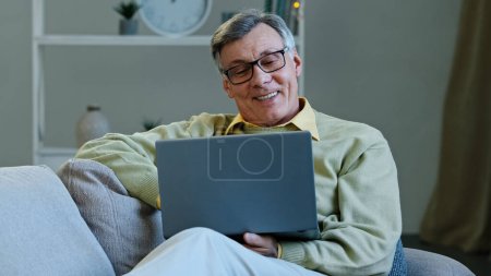 Photo for Happy grandfather sit on sofa living room mature elderly caucasian man looking at screen laptop work remotely browses news on internet make online purchases older people and modern technology concept - Royalty Free Image
