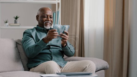 Foto de Happy old bald overweight African American senior mature elderly man happy with financial savings salary at home sitting couch. Winning online lottery holding dollars throw up cash money. High quality - Imagen libre de derechos