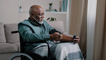 Photo for Side view African old 60s male ill senior retired bald man with gray beard measuring blood pressure with electronic medical device and stethoscope sitting in wheelchair at home cardiology healthcare - Royalty Free Image