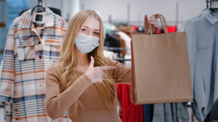 Photo for Close-up happy Caucasian woman in medical mask covid pandemic shopping stand in clothing store shop girl consumer showing pointing on bags purchases lady satisfied low prices discount Black Friday - Royalty Free Image
