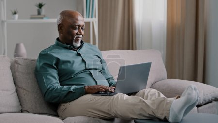 Foto de African bald overweight obese old middle-aged man senior businessman sitting on sofa at home working with laptop typing feels tired stress relaxing leaning back on sofa pauses after finishing work - Imagen libre de derechos