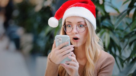 Photo for Surprised wonder astonish shocked amazed Caucasian woman girl lady in Santa Claus Christmas hat and eyeglass looking at phone mobile win victory offer achievement New Year celebration winning discount - Royalty Free Image