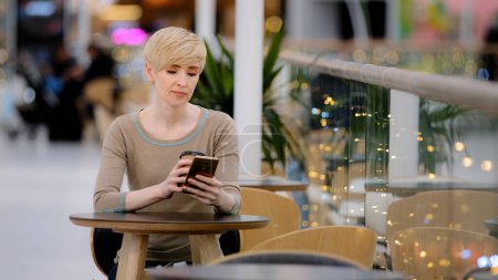 Foto de Middle-aged adult 40s caucasian woman businesswoman sitting at cafe table drinking coffee tea looking in mobile phone typing smartphone chatting browsing buying online app using wi-fi scrolling media - Imagen libre de derechos