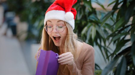 Téléchargez les photos : Fille caucasienne en chapeau Santa regarde dans le sac cadeau shopping violet ouvre paquet de carton femme heureuse souriante se réjouit d'acheter la danse heureuse avec cadeau intérieur millénial femme joyeusement célébrer - en image libre de droit