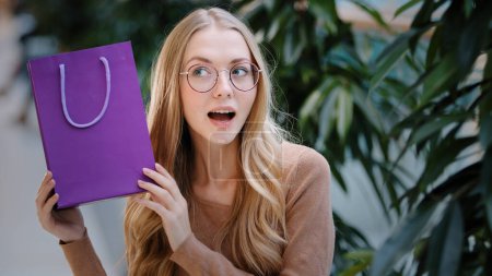 Photo for Portrait excited young woman with glasses shakes gift bag amazing caucasian millennial birthday girl opens purple package feels joy satisfaction blonde female smiling happily dancing enjoying surprise - Royalty Free Image