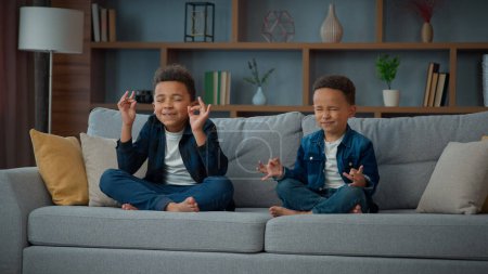 Téléchargez les photos : Two funny little African American ethnic children boys brothers siblings kids meditating together sitting on couch in lotus position. Small schoolboys meditate. Spiritual development yoga meditation - en image libre de droit