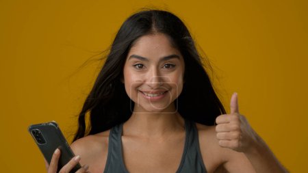 Photo for Portrait Indian ethnic girl woman female lady with mobile phone win victory winning showing thumb up achieve success recommend smartphone app online web chat super gesture in studio yellow background - Royalty Free Image