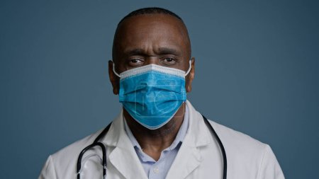 Téléchargez les photos : Serious african american mature man doctor surgeon therapist in medical gown and protective mask looking at camera posing in studio gray background medical service healthcare health protection concept - en image libre de droit