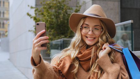 Foto de Caucasian girl blogger shoot video for blog with smartphone after shopping in city woman taking photo on mobile camera millennial lady female buyer client has video call conference with phone outdoors - Imagen libre de derechos