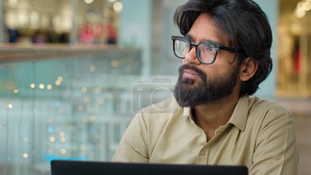 Photo for Concentrated pensive thoughtful Indian Arabian ethnic multiracial bearded businessman trader man entrepreneur male manager CEO in eyeglasses with laptop indoor looking to side thinking pondering think - Royalty Free Image