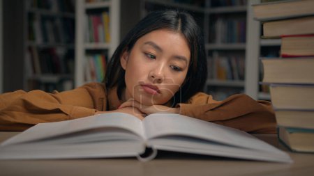 Gelangweilt traurig müde faule junge Koreanerin lesen langweilige Buch auf dem Schreibtisch liegend in der Universitätsbibliothek unmotivierte Studentin Hausaufgaben machen Vorbereitung auf College-Prüfung Überlastung mit Studium liebloses Thema