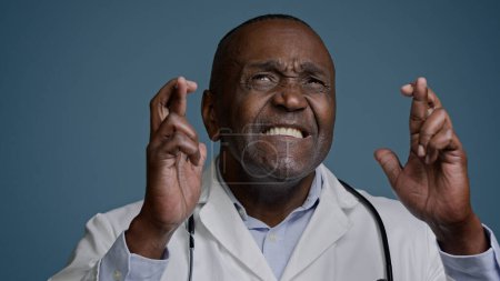 Téléchargez les photos : Close-up mature man in studio on gray background hopeful doctor surgeon crosses fingers for good luck makes wish dreaming praying for success asks for help believe in victory waiting for great results - en image libre de droit