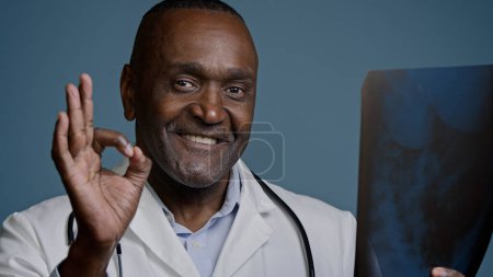 Photo for Close-up mature african american man doctor surgeon traumatologist radiologist in medical coat in studio blue background hold x-ray review result tomogram showing sign ok gesture approval good health - Royalty Free Image