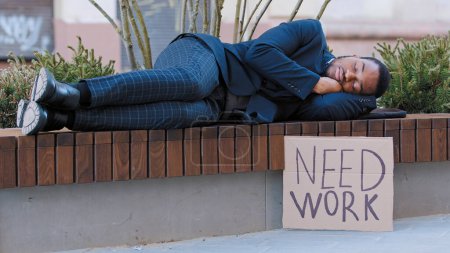 Foto de Sleeping homeless fired African American man in city tired with searching job relax on bench. Male sleep outdoors with poster need work. Illegal ethnic guy lost job poor jobless unemployment racism - Imagen libre de derechos