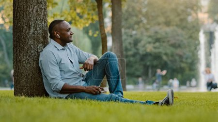 Téléchargez les photos : Heureux calme détendu âgé d'âge moyen écoute de musique masculine avec écouteurs mature adulte afro-américain homme ethnique dans des écouteurs sans fil assis sur l'herbe dans le parc de la ville profiter du son de la chanson audio - en image libre de droit