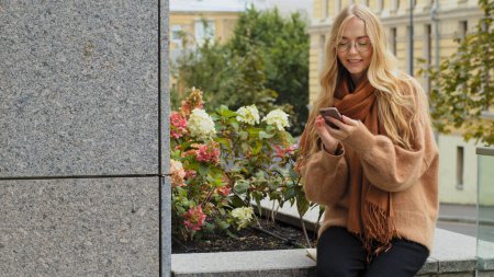 Foto de Caucasian woman browsing mobile phone girl in glasses sitting outdoors in city scrolling smartphone chat female lady businesswoman reading news in cellphone online app chatting texting in net outside - Imagen libre de derechos