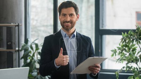 Foto de Focused adult caucasian man recruiting agent standing in office making notes writing checklist businessman conducts inspection write strategic plan showing thumb up gesture approval best choice sign - Imagen libre de derechos