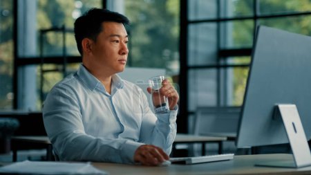 Téléchargez les photos : Asiatique coréen homme boire verre d'eau douce froide garder équilibre santé rajeunissement énergie santé habitude homme d'affaires assis au bureau travailler sur ordinateur faire pause étancher soif corps hydratation soins du corps - en image libre de droit