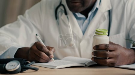 Photo for Unknown male doctor african american ethnic man sit at desk with stethoscope write notes prescribe medical insurance checklist writing dosage of medication hold bottle drug antibiotic pills vitamins - Royalty Free Image