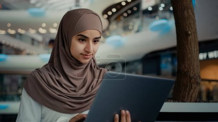 Téléchargez les photos : Arabe islamique musulmane femme d'affaires dans hijab femme entrepreneure étudiante femme travaillant au centre commercial utiliser ordinateur portable navigation internet recherche emploi vacance faire l'ordre achat apprentissage en ligne distant - en image libre de droit