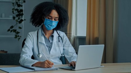 Foto de Mujer africana étnica médico terapeuta cardiólogo psicoterapeuta en máscara médica escribir diagnóstico de prescripción hablando en video chat llamando paciente distante tratamiento en línea durante covid-19 epidemia - Imagen libre de derechos