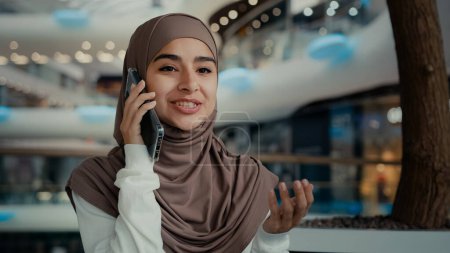 Téléchargez les photos : Belle femme musulmane dans le hijab assis dans un lieu public parler téléphone mobile au centre commercial femme millénariste arabe fille détendue femme d'affaires appelant éloigné pour discuter conversation d'affaires - en image libre de droit