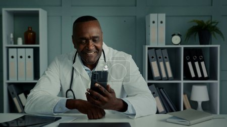 Téléchargez les photos : Médecin africain homme d'âge moyen travailleur utiliser téléphone portable gadget texto message bavarder clients de consultation en ligne via application mobile de soins de santé assis dans la clinique de bureau technologie moderne et de soins de santé - en image libre de droit