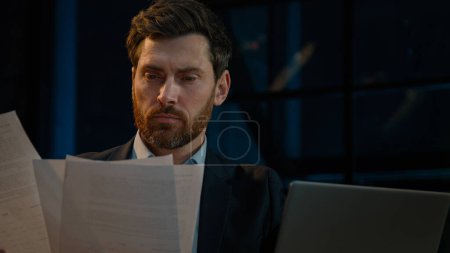 Foto de Caucasian 40s businessman employee auditor middle aged man sit in evening night office read business documents papers legal contracts pensive employee think check data in computer search information - Imagen libre de derechos