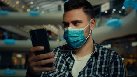 Photo for Businessman in medical mask caucasian man chatting online on smartphone scrolling internet website use mobile phone sick male protect face from coronavirus infection quarantine rules in shopping mall - Royalty Free Image