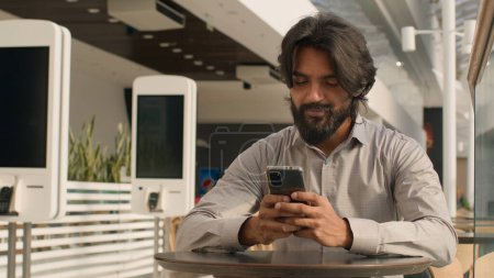 Téléchargez les photos : Happy young indian businessman holding smartphone sending message sitting indoors smiling arabian man play mobile game using application on phone reading good news on internet chatting in social media - en image libre de droit