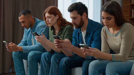 Photo for Millennial 30s diverse people sit in line on sofa holding smartphone ignoring friends chatting online in messenger mobile phone addicted women men internet overuse problem modern wireless tech usage - Royalty Free Image