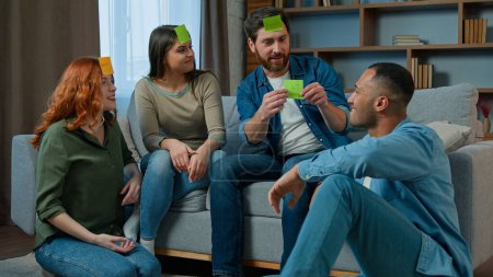 Foto de Diverse interracial friends women men playing guessing game with sticky notes on forehead play charades asking questions funny entertainment at home african american ethnic man winner win gaming level - Imagen libre de derechos