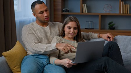 Foto de African american latin couple look at laptop multiracial woman and man rest on couch diverse family partners watching film talking use computer device choosing e-services shopping online browsing news - Imagen libre de derechos