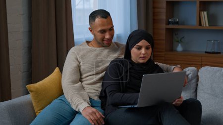 Foto de Married couple sit on sofa use laptop device choosing new apartment on internet website multiracial family shocked amazed diverse man woman search information by computer read good news talking indoor - Imagen libre de derechos