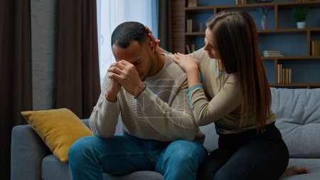 Foto de Offended African american man sit on couch upset with bad news health problem Caucasian woman consoling apologize ask forgiveness after quarrel wife touching husband calming support family conflict - Imagen libre de derechos