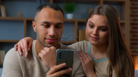 Photo for Ethnic couple use mobile phone apps smiling african american man and caucasian woman looking at smartphone screen watch video online social media chatting home leisure with cellphone internet shopping - Royalty Free Image