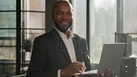 Foto de Senior mature businessman African American ethnic man male worker develop business project online on laptop computer typing on keyboard stand in sunny sun rays office agency looking at camera smiling - Imagen libre de derechos