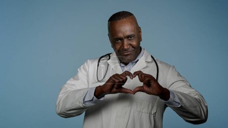 Photo for Mature smiling african american man doctor cardiologist in white coat stand in blue background studio showing shape heart gesture love support cardiology services charity medical insurance healthcare - Royalty Free Image