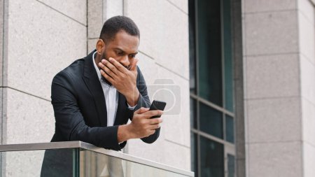 Téléchargez les photos : Frustré ethnique afro-américain barbu triste homme choqué regardant l'écran du smartphone se sentir déçu par reçu de mauvaises nouvelles message stress homme d'affaires défaillance de téléphone mobile debout sur le balcon - en image libre de droit