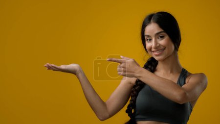 Téléchargez les photos : Portrait en studio Indien 20s femme posant sur fond jaune pointant vers le côté au bras de la paume pointe de côté recommander signe fille dame montrant à vide espace de copie en regardant la caméra - en image libre de droit