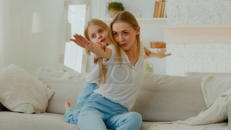 Téléchargez les photos : Caucasian mother with little daughter piggyback pretending flying stretch hands to side playing airplane game sitting at couch in living room. Mom with child play kid girl having fun at sofa at home - en image libre de droit