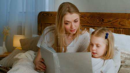 Foto de Family learning concept. Caucasian mother help little small cute daughter preschool child girl baby reading book in bed at home bedroom mom with kid read literature fairy tale elementary education - Imagen libre de derechos