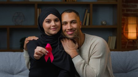 Téléchargez les photos : Multiracial family muslim woman and african american man sit on sofa indoors hold red ribbon prevent symbol HIV AIDS protect immune deficiency syndrome blood donation health care and world cancer day - en image libre de droit