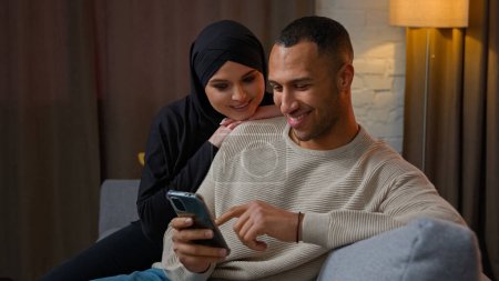Téléchargez les photos : Famille couple multiethnique interracial famille afro-américain homme bavarder sur téléphone portable femme musulmane câlin mari utiliser téléphone portable shopping en ligne ensemble regarder drôle vidéo internet à la maison - en image libre de droit