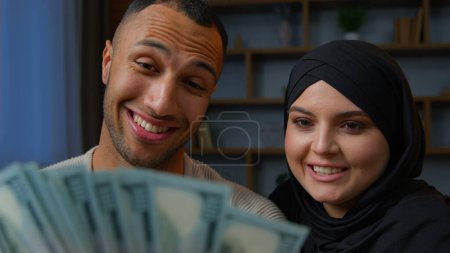Foto de Rich multiracial couple spouses African American man counting money dollars with Arabian muslim woman in hijab planning family budget diverse partners considers finances cash banknotes sitting on sofa - Imagen libre de derechos