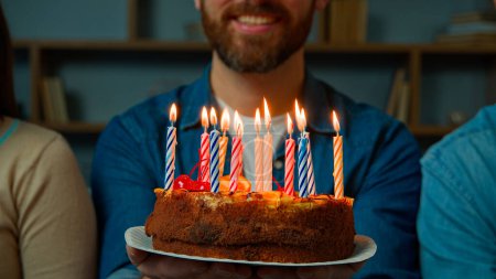 Téléchargez les photos : Gros plan gâteau festif avec des bougies allumées heureux barbu méconnaissable anniversaire homme souriant faire vœu célébrer événement avec des amis à la maison fête anniversaire année tradition félicitations - en image libre de droit